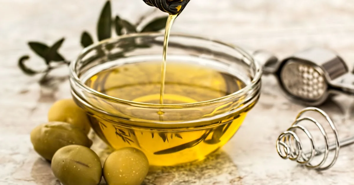 Gros plan sur de l'huile d'olive versée dans un bol en verre entouré d'olives fraîches et d'ustensiles de cuisine.