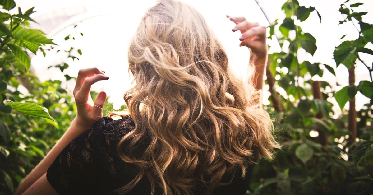 Femme blonde aux cheveux bouclés, entourée de verdure luxuriante, profitant d'une journée ensoleillée.