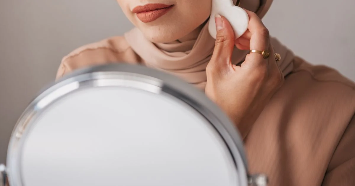 Gros plan d'une femme en hijab s'appliquant du maquillage avec une éponge faciale devant un miroir.