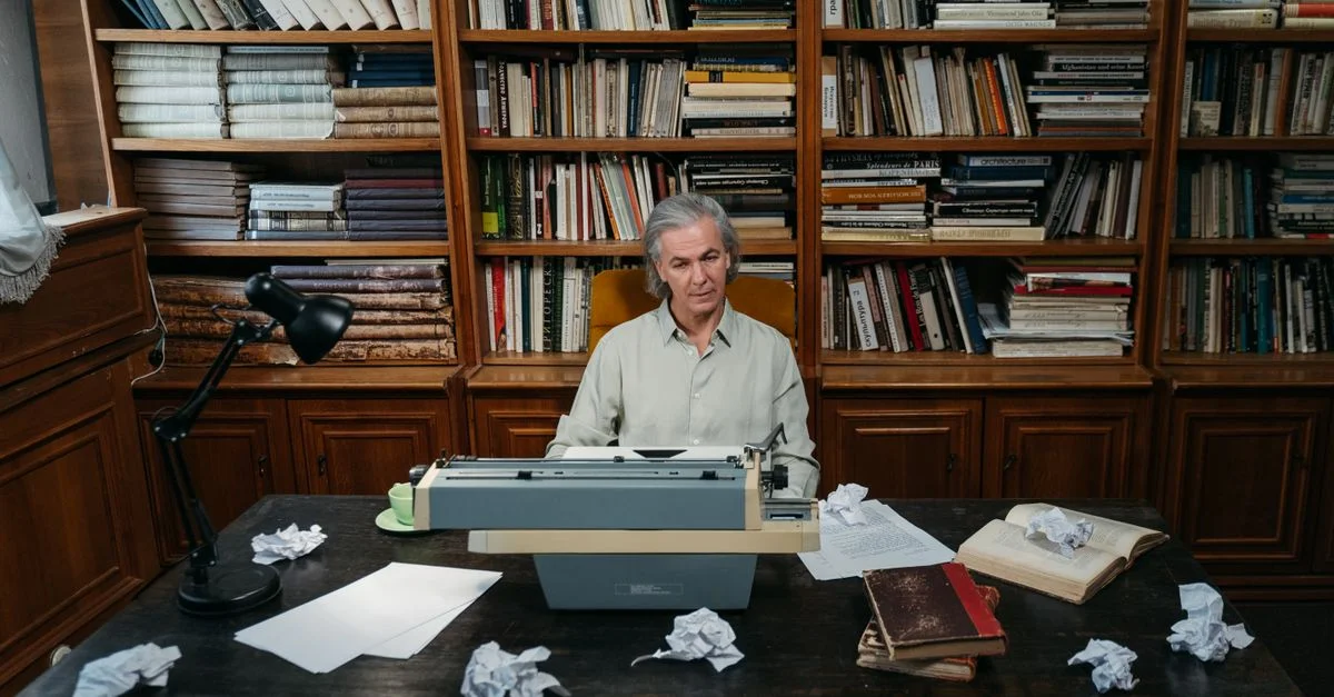 Un homme âgé à une machine à écrire dans une bibliothèque, entouré de livres et de papier.