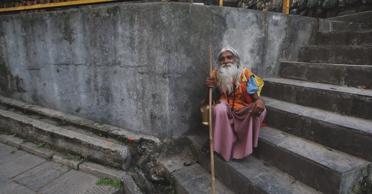 Un moine âgé et contemplatif avec une canne repose sur des escaliers en pierre près des roues de prière.