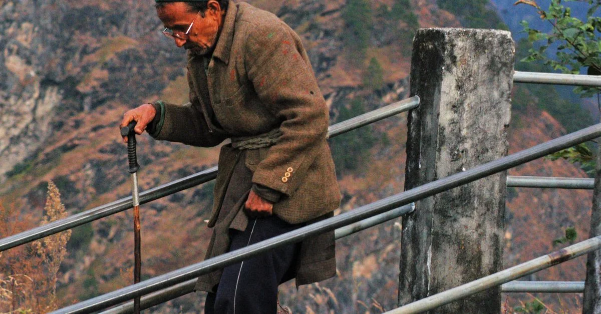 Un homme âgé utilisant une canne monte une colline sur un sentier de montagne pittoresque.